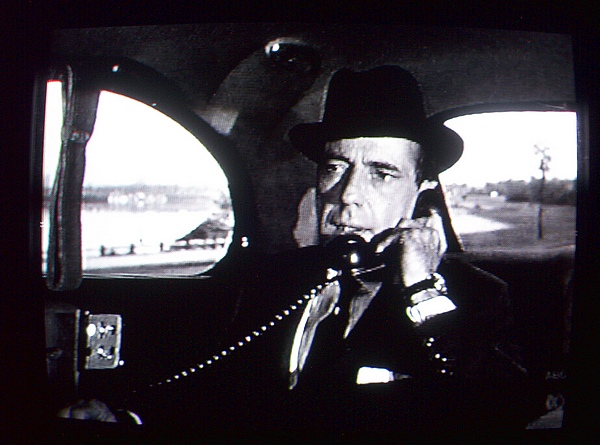 Humphrey Bogart sitting in the back of a car, wearing a suit and dark hat. He is holding a 1940s telephone to his ear